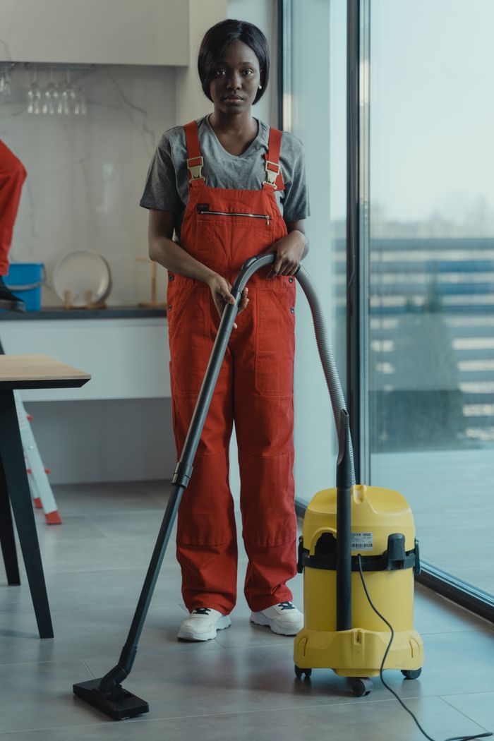 hero-img-01 An adult woman in red coveralls operates a vacuum cleaner in a modern indoor setting, focusing on cleaning tasks.